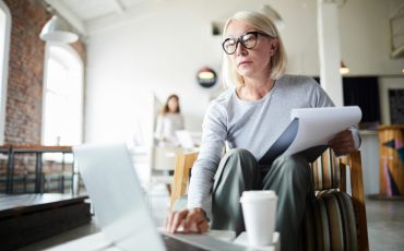 Mature businesswoman with document and laptop sitting on chair in office and browsing in the net